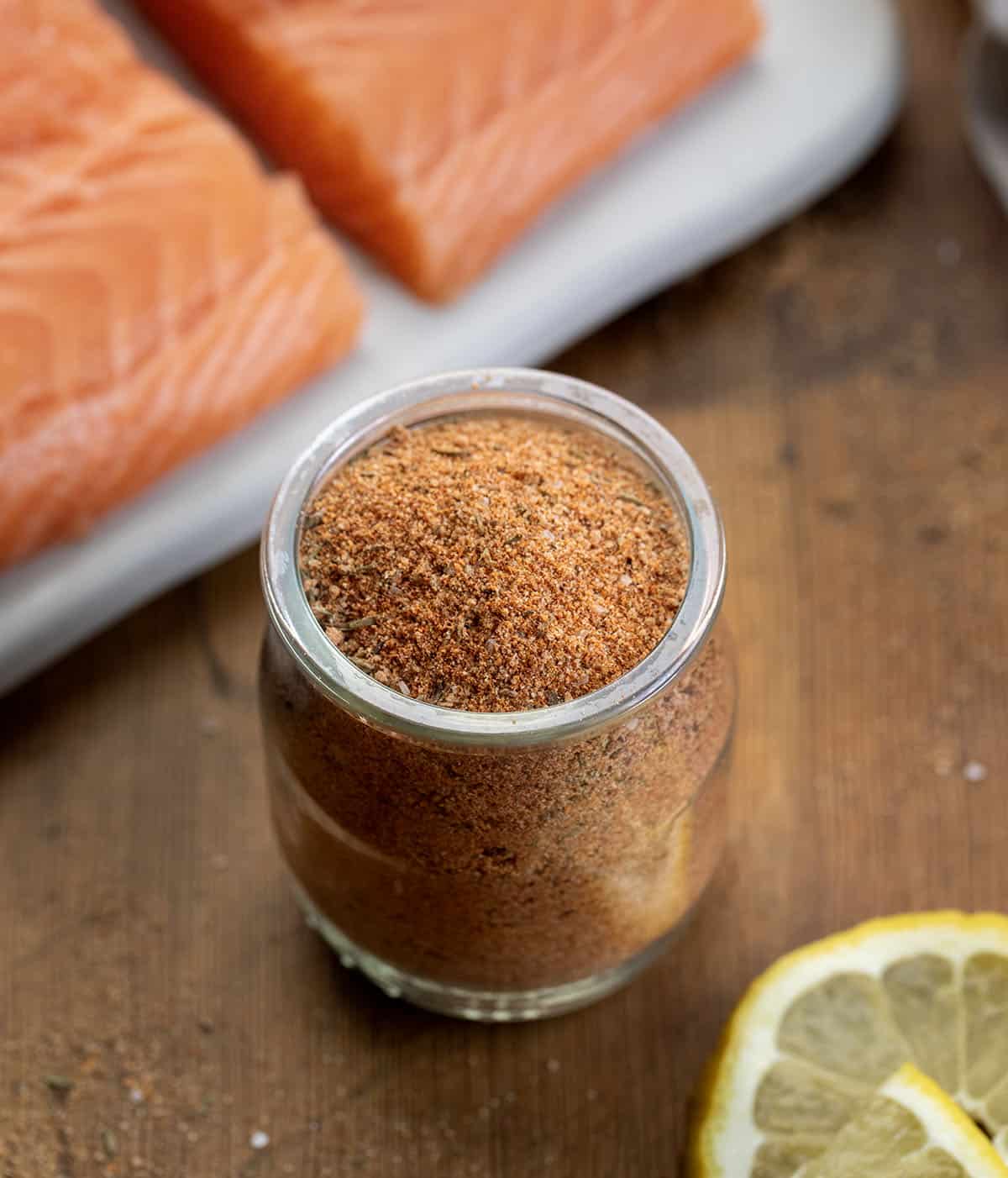 Glass jar of Salmon Seasoning on a wooden table next to fresh salmon and lemon slices Glass jar of Salmon Seasoning on a wooden table next to fresh salmon and lemon slices.
