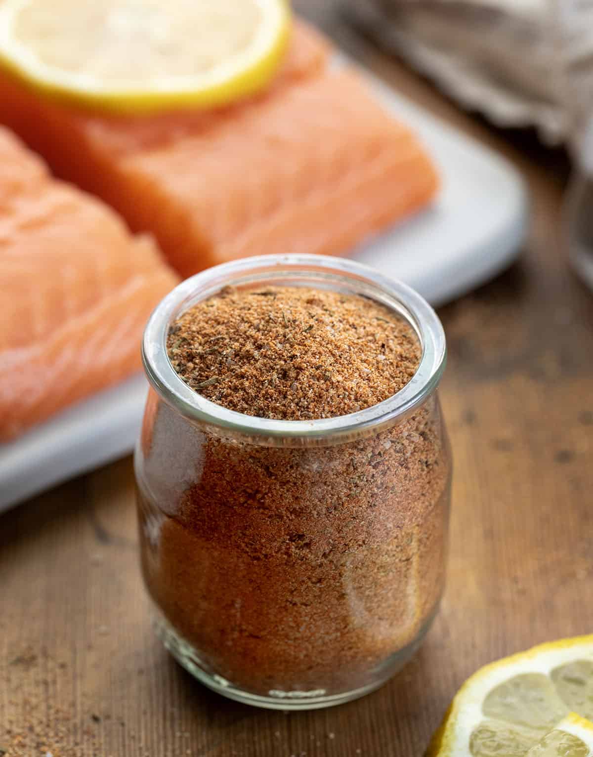 Glass jar of Salmon Seasoning on a wooden table next to fresh salmon and lemon slices Glass jar of Salmon Seasoning on a wooden table next to fresh salmon and lemon slices.