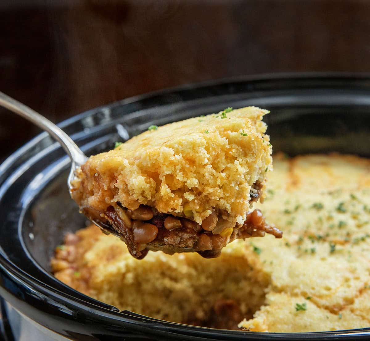 Scoop of Slow Cooker Cornbread Cowboy Casserole being held above the slow cooker Scoop of Slow Cooker Cornbread Cowboy Casserole being held above the slow cooker.