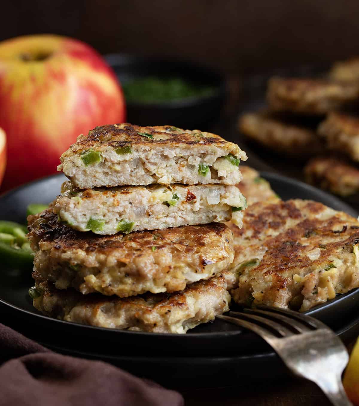 One Apple Jalapeño Breakfast Sausage Patty cut in half sitting on a plate with more patties on it near apples and jalapenos One Apple Jalapeño Breakfast Sausage Patty cut in half sitting on a plate with more patties on it near apples and jalapenos.