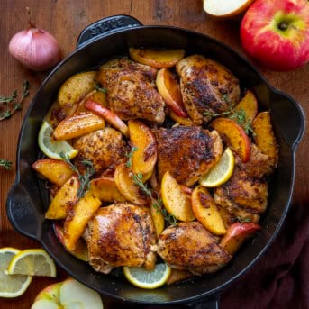 Skillet of Apple Shallot Chicken on a wooden table with fresh shallot, apples, and lemon.