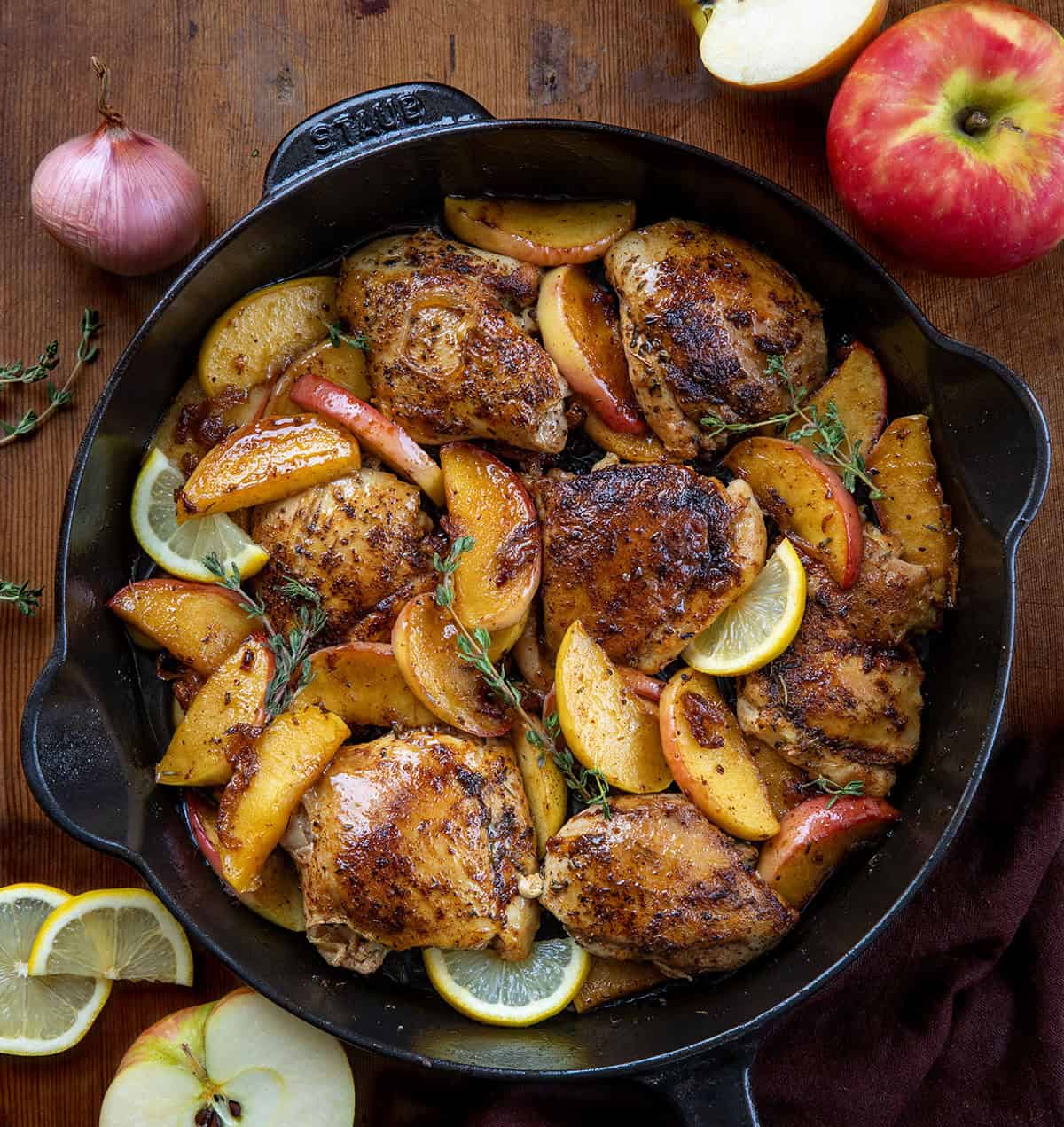 Skillet of Apple Shallot Chicken on a wooden table with fresh shallot, apples, and lemon Skillet of Apple Shallot Chicken on a wooden table with fresh shallot, apples, and lemon.