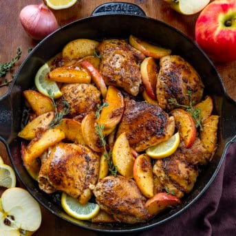 Skillet of Apple Shallot Chicken on a wooden table with fresh shallot, apples, and lemon.
