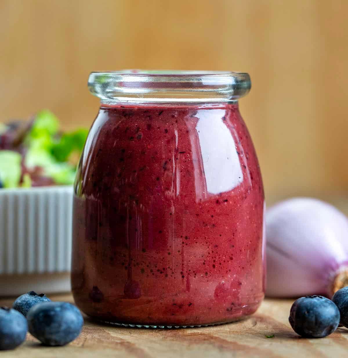 Jar of Blueberry Balsamic Vinaigrette on a wooden table with salad in the background Jar of Blueberry Balsamic Vinaigrette on a wooden table with salad in the background.