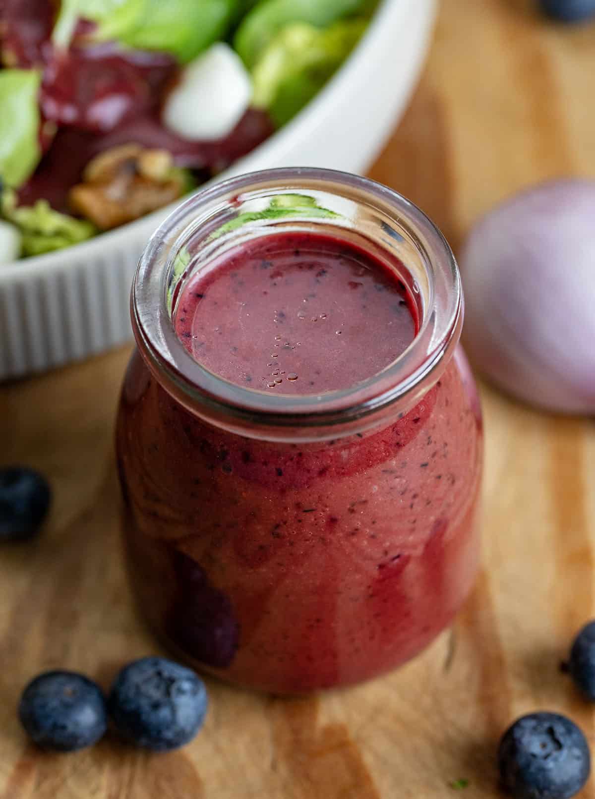 Jar of Blueberry Balsamic Vinaigrette on a wooden table with salad, shallot, and blueberries Jar of Blueberry Balsamic Vinaigrette on a wooden table with salad, shallot, and blueberries.