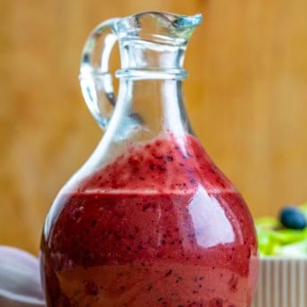 Glass jar of Blueberry Balsamic Vinaigrette on a wooden table with salad and fresh blueberries.