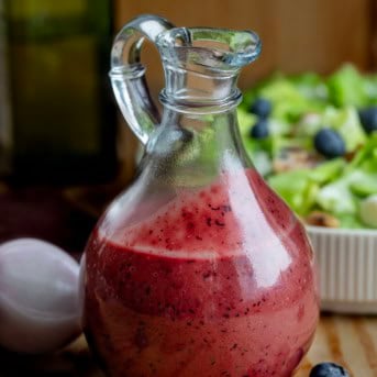 Glass jar of Blueberry Balsamic Vinaigrette on a wooden table with salad, olive oil, shallot, and blueberries.