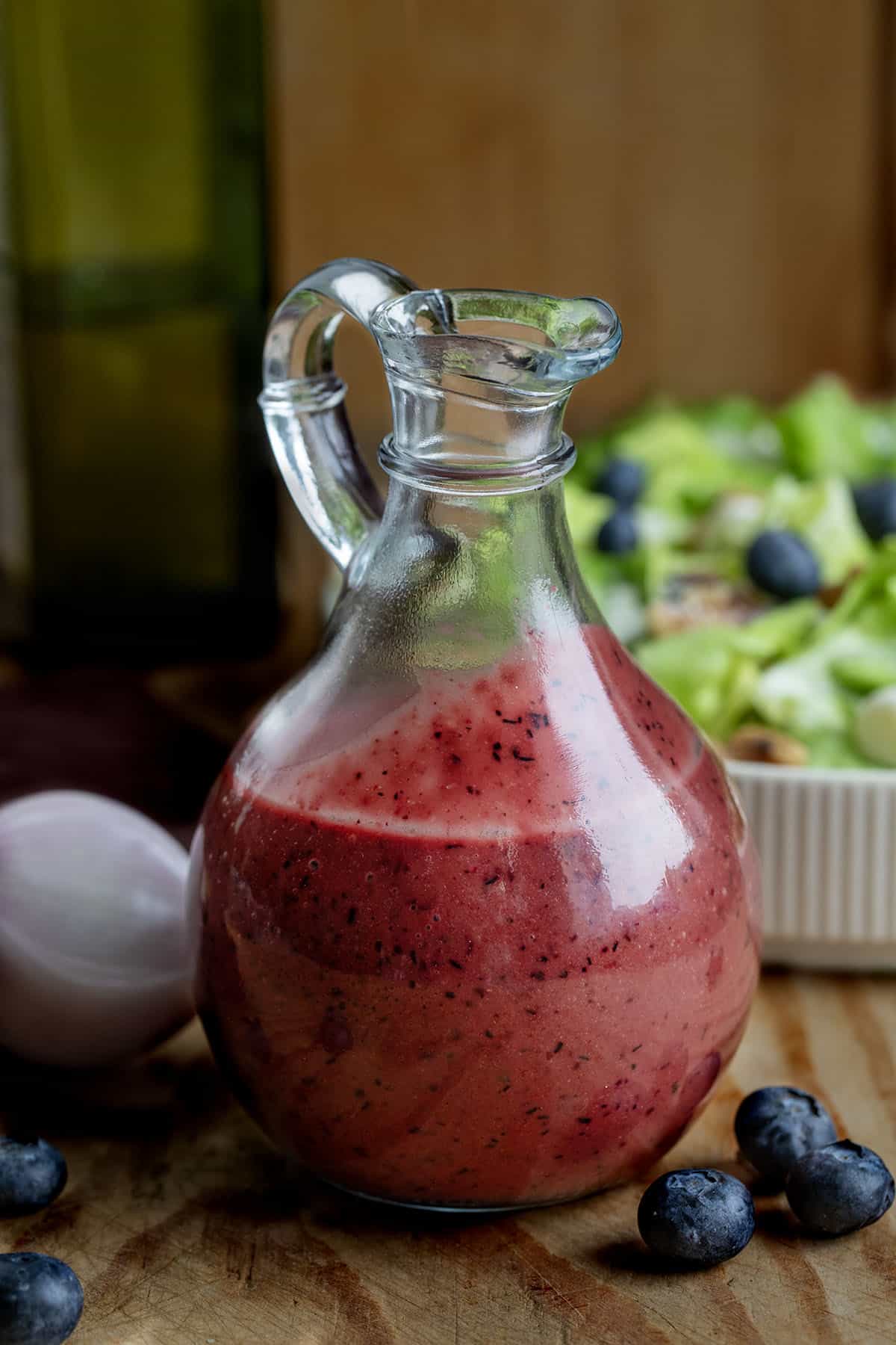 Glass jar of Blueberry Balsamic Vinaigrette on a wooden table with salad, olive oil, shallot, and blueberries Glass jar of Blueberry Balsamic Vinaigrette on a wooden table with salad, olive oil, shallot, and blueberries.