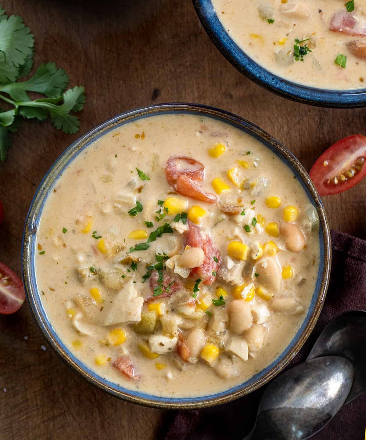 Pot of Creamy White Bean Chicken Chili on a wooden table surrounded by bowls and cilantro and tomatoes Creamy White Bean Chicken Chili in a blue bowl on a wooden table from overhead.