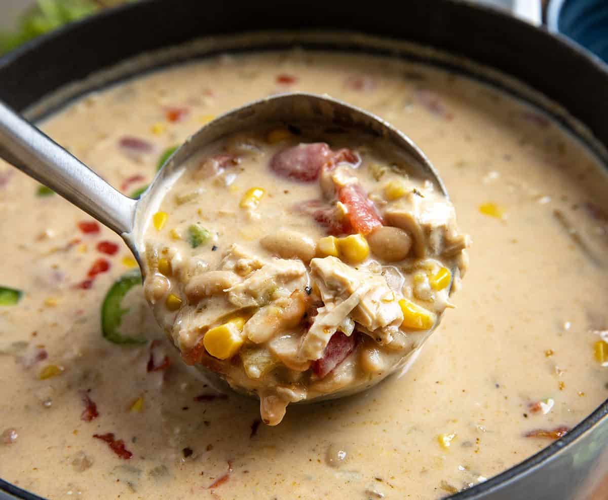 Pot of Creamy White Bean Chicken Chili on a wooden table surrounded by bowls and cilantro and tomatoes Ladle full of Creamy White Bean Chicken Chili being held above the pot.