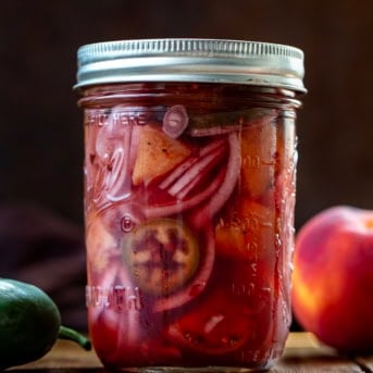 Chilled jar of Jalapeño Peach Pickled Onions on a wooden table with fresh jalapeno and peach.