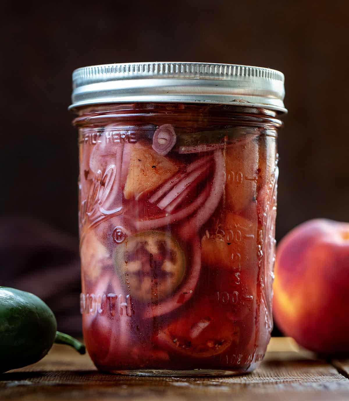 Chilled jar of Jalapeño Peach Pickled Onions on a wooden table with fresh jalapeno and peach Chilled jar of Jalapeño Peach Pickled Onions on a wooden table with fresh jalapeno and peach.