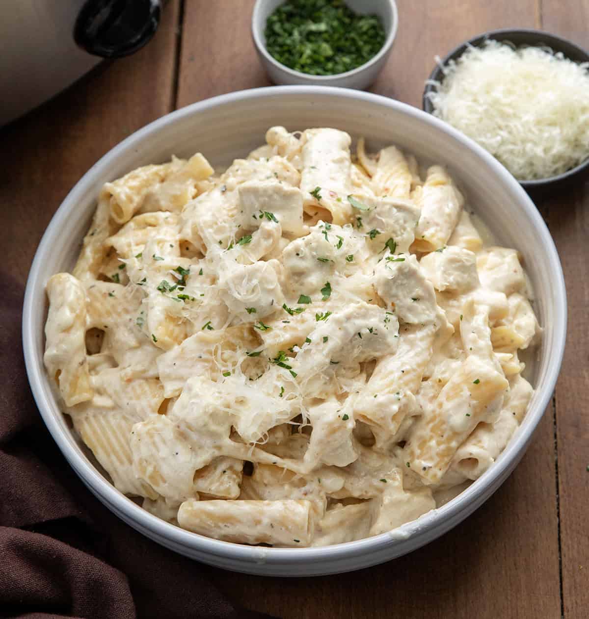 Bowl of Slow Cooker Chicken Alfredo Pasta next to the slow cooker on a wooden table Bowl of Slow Cooker Chicken Alfredo Pasta next to the slow cooker on a wooden table.