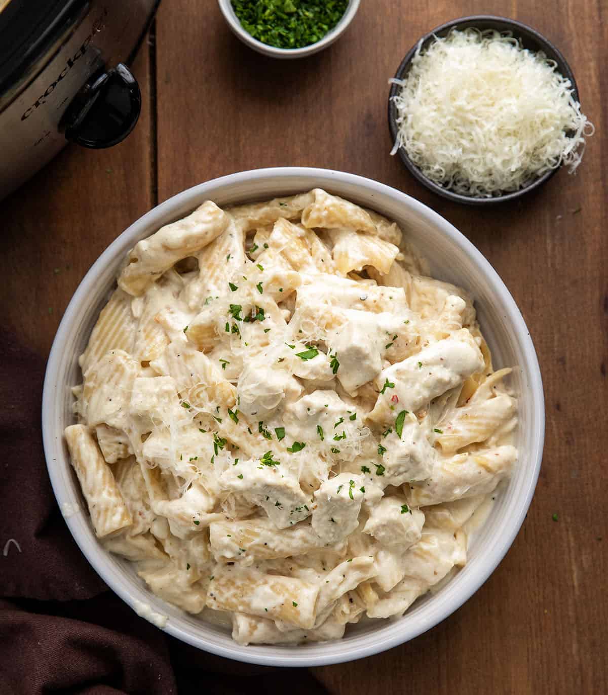 Bowl of Slow Cooker Chicken Alfredo Pasta next to the slow cooker on a wooden table Bowl of Slow Cooker Chicken Alfredo Pasta next to the slow cooker on a wooden table.