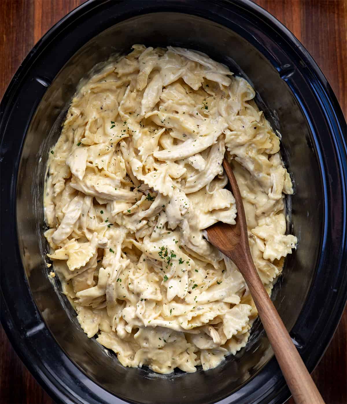 Slow Cooker Chicken Alfredo Pasta with a wooden spoon in it on a wooden table Slow Cooker Chicken Alfredo Pasta with a wooden spoon in it on a wooden table.