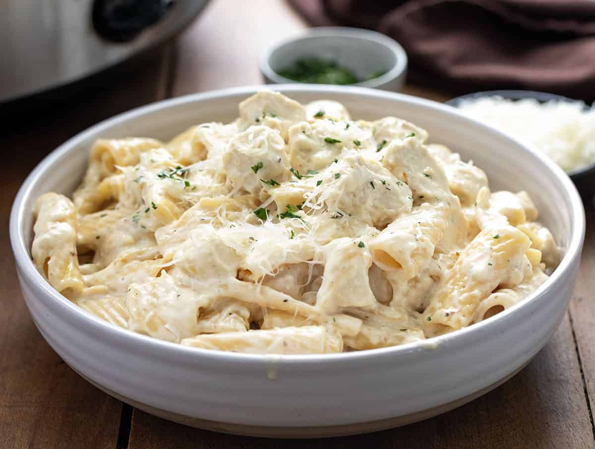 Bowl of Slow Cooker Chicken Alfredo Pasta next to the crockpot on a wooden table Bowl of Slow Cooker Chicken Alfredo Pasta next to the crockpot on a wooden table.
