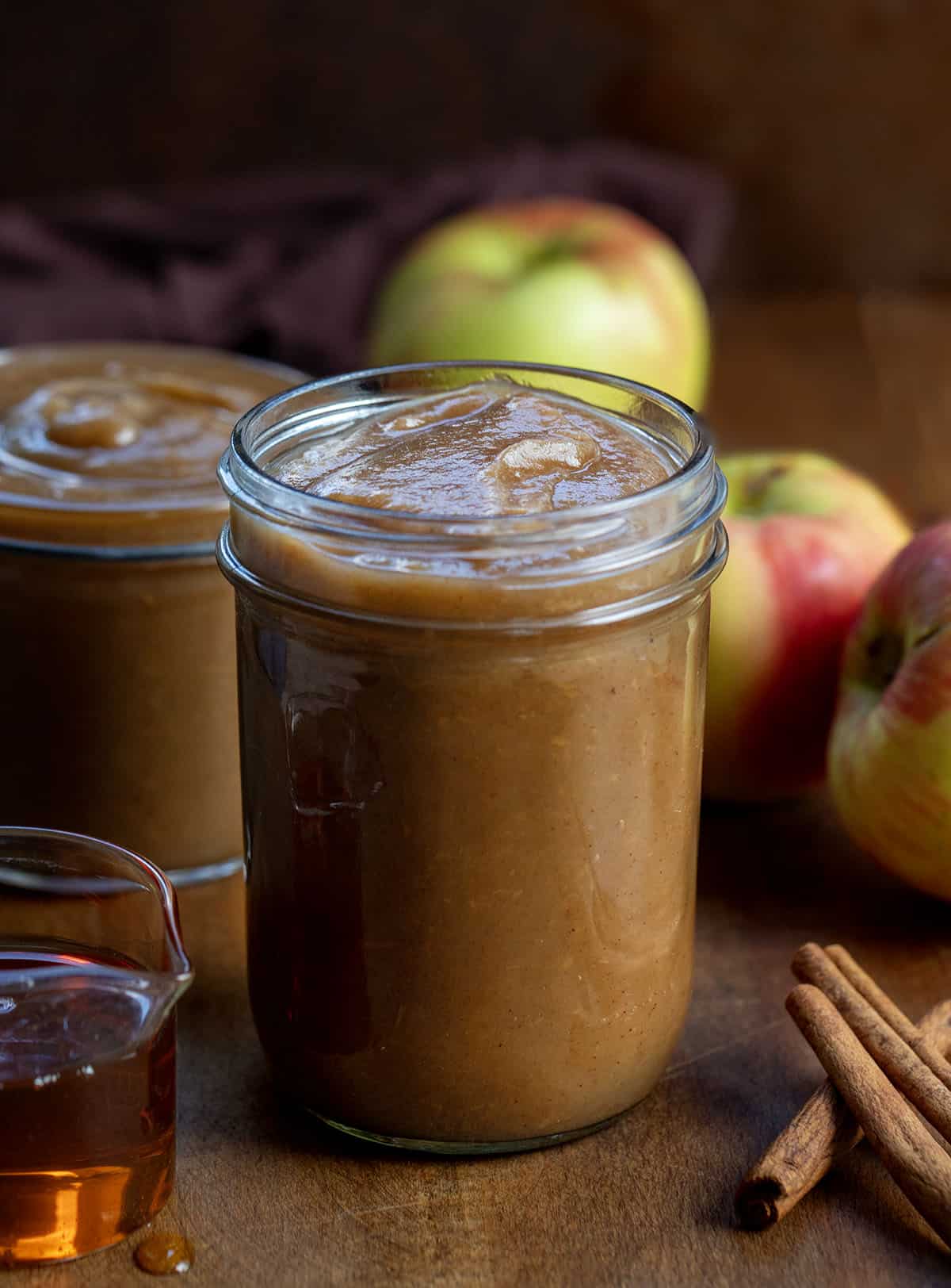 Jars of Slow Cooker Maple Apple Butter on a wooden table with apples, cinnamon, and maple syrup Jars of Slow Cooker Maple Apple Butter on a wooden table with apples, cinnamon, and maple syrup.