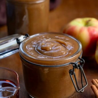 Jar of Maple Apple Butter on a wooden table with apples and cinnamon.