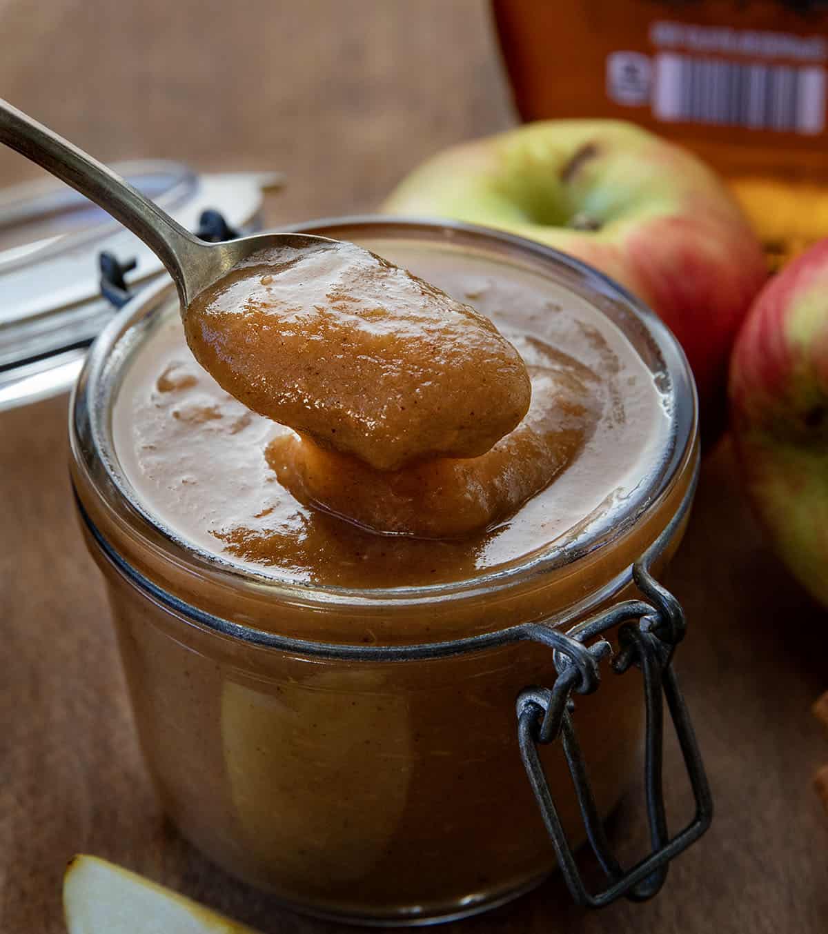 Spoonful of Maple Apple Butter being held above the jar on a wooden table with apples and maple syrup Spoonful of Maple Apple Butter being held above the jar on a wooden table with apples and maple syrup.