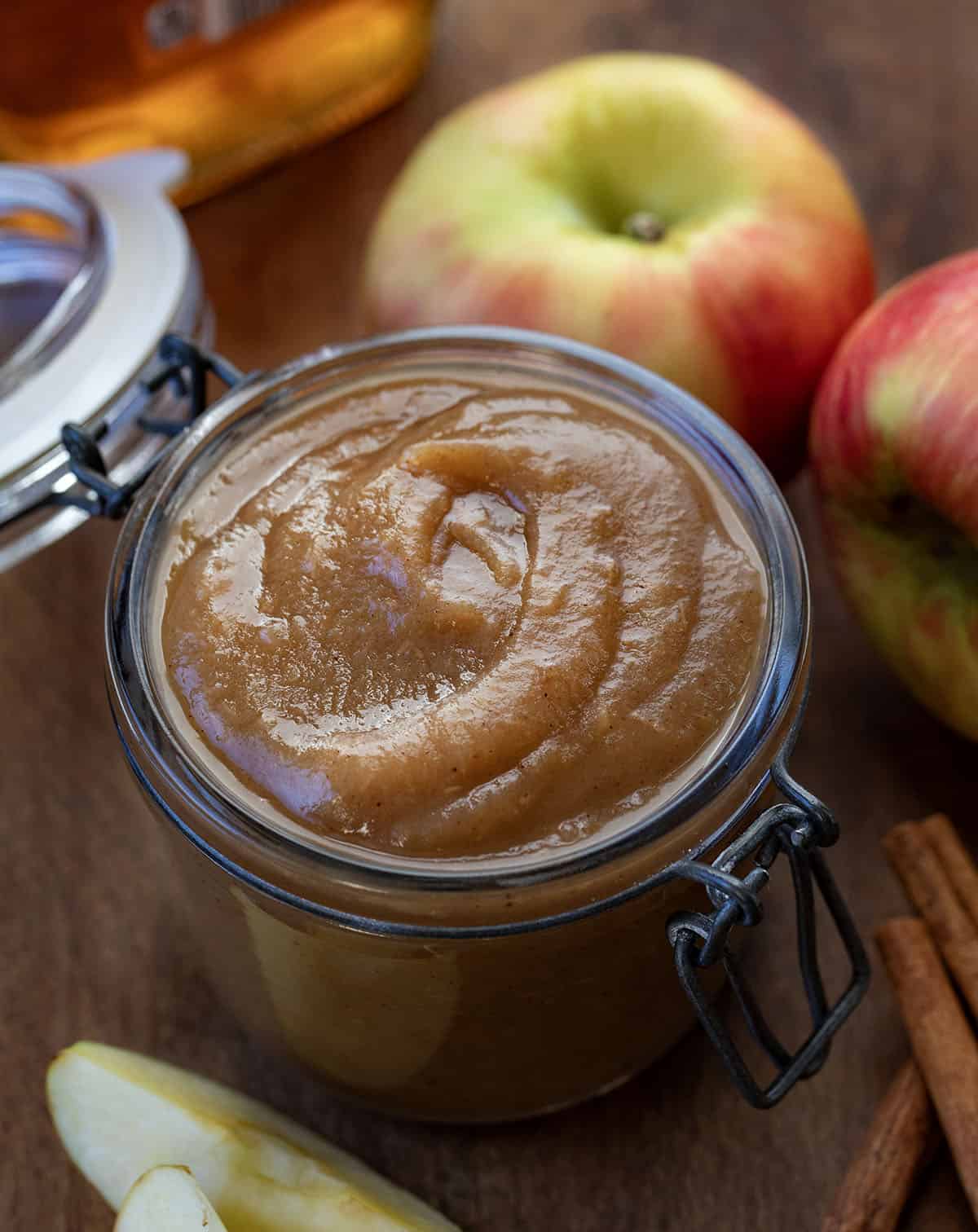 Jar of Maple Apple Butter on a wooden table with apples, cinnamon sticks, and maple syrup Jar of Maple Apple Butter on a wooden table with apples, cinnamon sticks, and maple syrup.