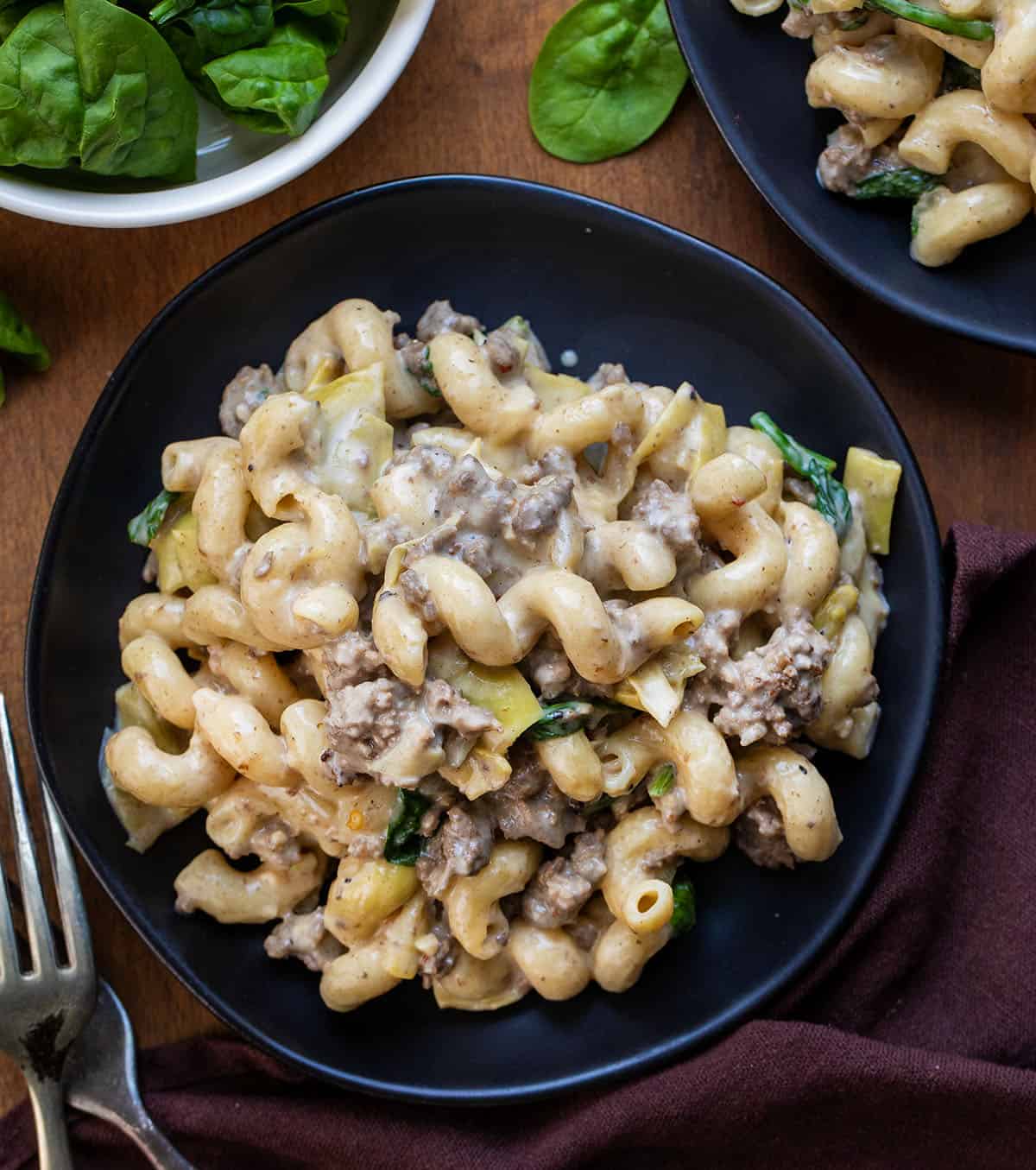 Black plates of Spinach Artichoke Hamburger Pasta on a wooden table from overhead Black plates of Spinach Artichoke Hamburger Pasta on a wooden table from overhead.