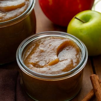 Jars of Stovetop Apple Butter on a wooden table with apples and cinnamon.