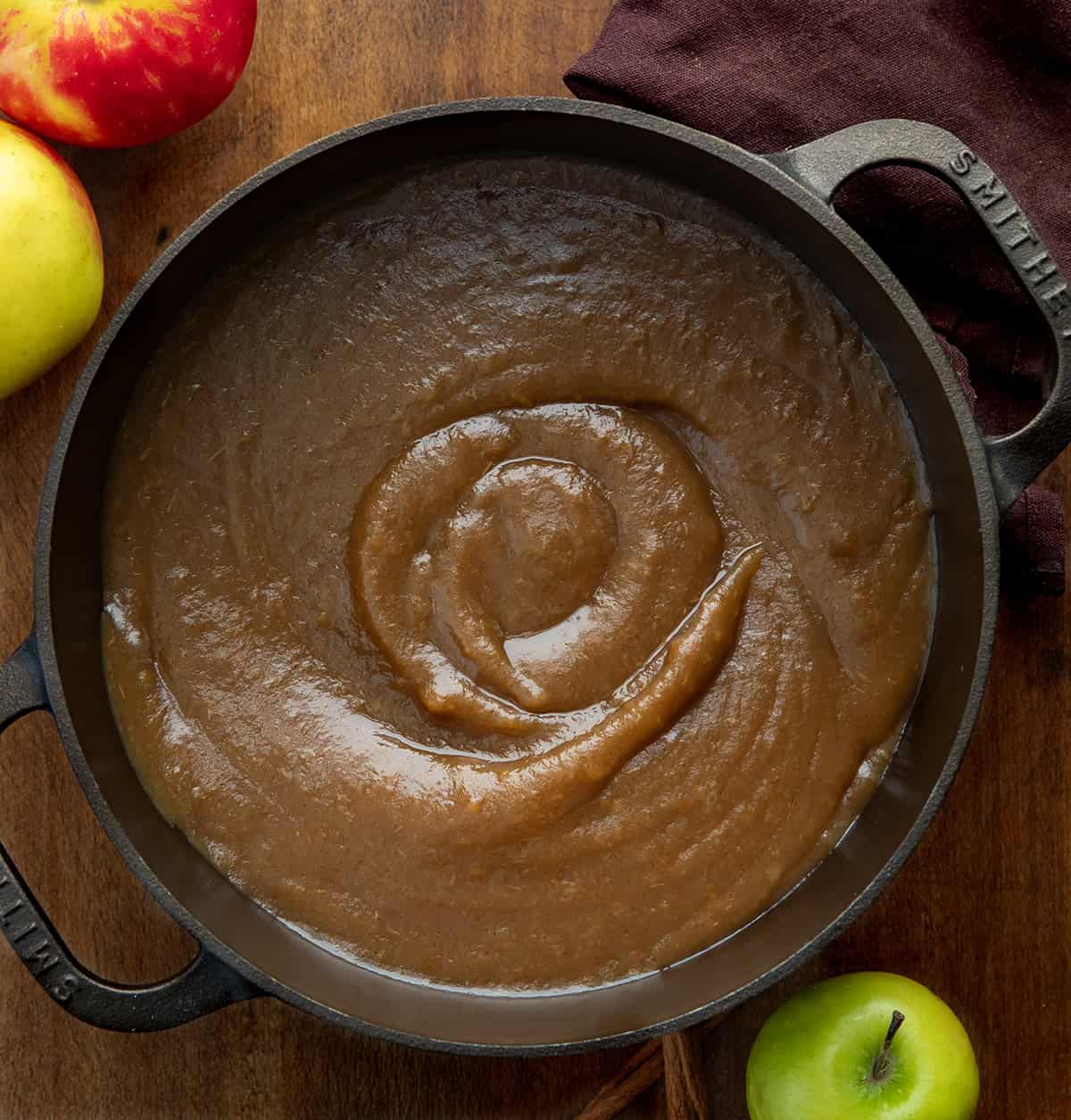 Pot of Stovetop Apple Butter on a wooden table with a brown napkin and apples from overhead Pot of Stovetop Apple Butter on a wooden table with a brown napkin and apples from overhead.