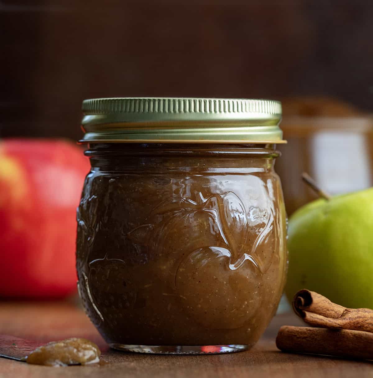 Sealed jar of Stovetop Apple Butter on a wooden table with apples behind it Sealed jar of Stovetop Apple Butter on a wooden table with apples behind it.