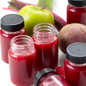 Small jars, some unopened, of Beet Raspberry Energy Juice on a white able with a beet, apple, and fresh raspberry.
