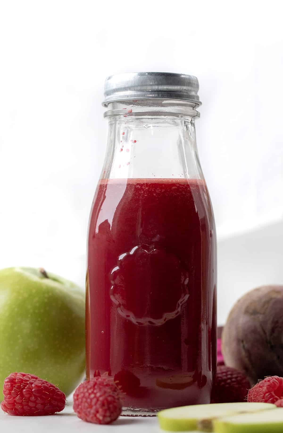 One jar of Beet Raspberry Energy Shot on a white table with apple, beet, and raspberry.