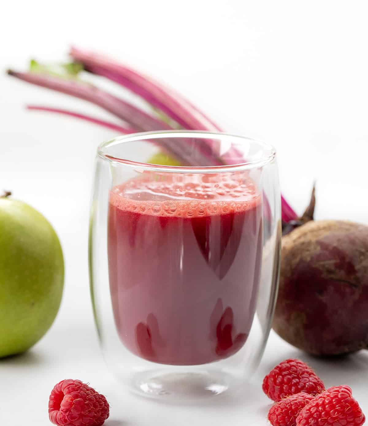 Glass of Beet Raspberry Energy Shot surrounded by beet, apple, and raspberry.