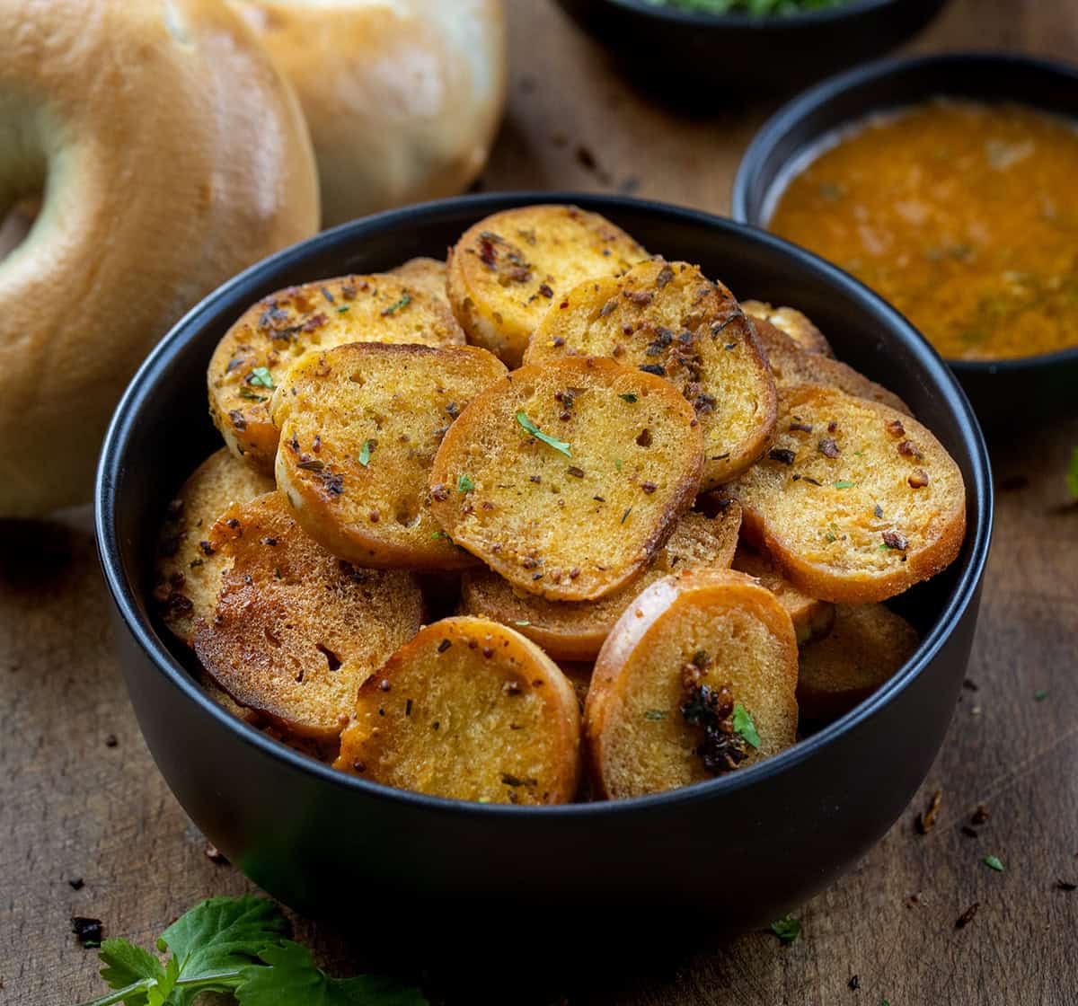 Black bowl of Cowboy Butter Bagel Chips on a wooden table with whole bagels and cowboy butter.