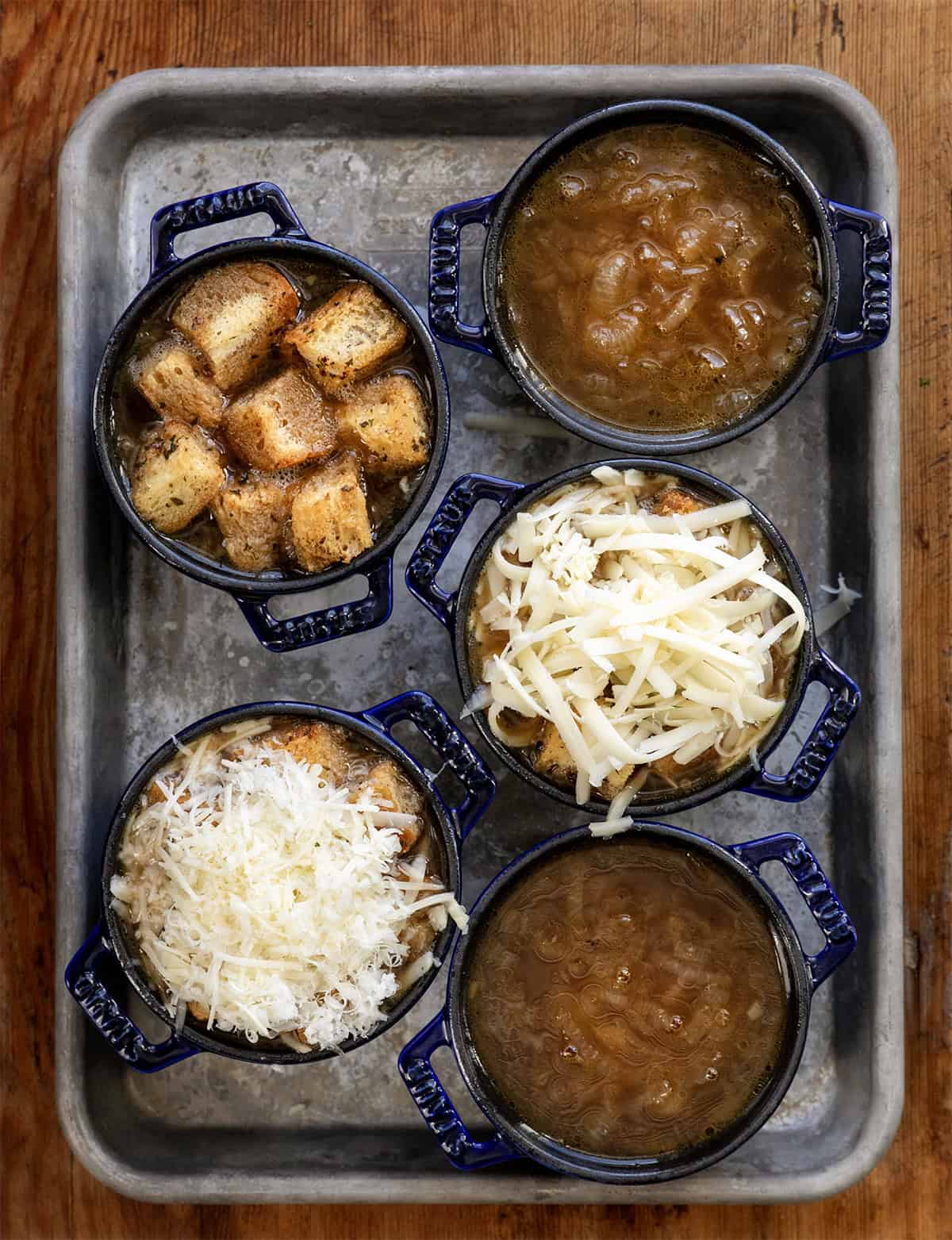 Small blue crocks of French Onion Soup ; some with soup, homemade croutons, gruyere, and parmesan cheese Small blue crocks of French Onion Soup ; some with soup, homemade croutons, gruyere, and parmesan cheese.