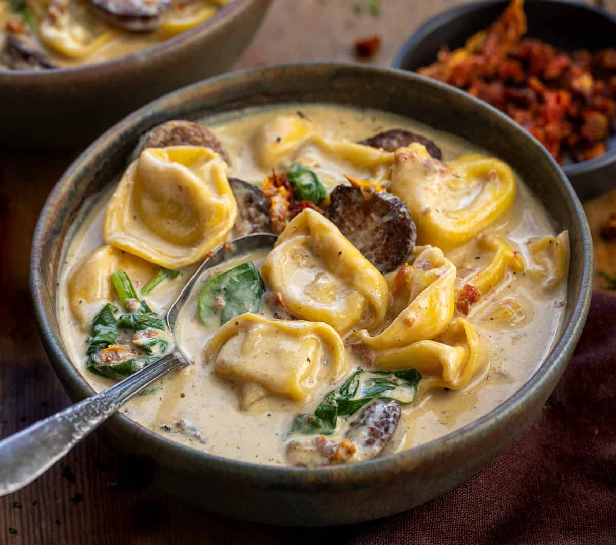 Bowl of Slow Cooker Kielbasa Tortellini Soup with a spoon in it on a wooden table Bowl of Slow Cooker Kielbasa Tortellini Soup with a spoon in it on a wooden table.
