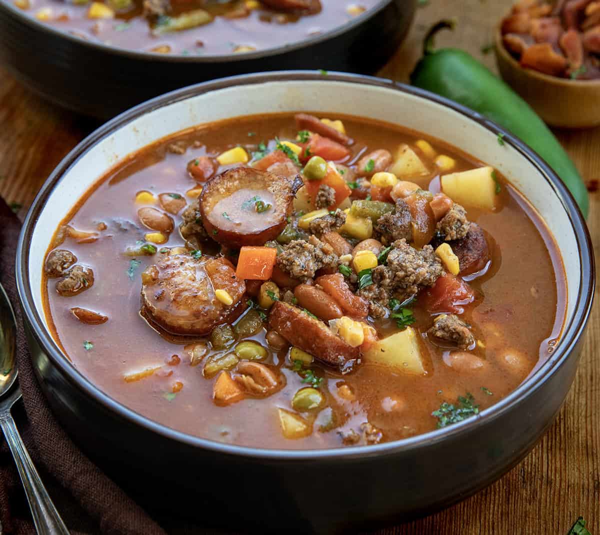 Bowl of Green Chili Cowboy Stew on a wooden table with another bowl behind it Bowl of Green Chili Cowboy Stew on a wooden table with another bowl behind it.
